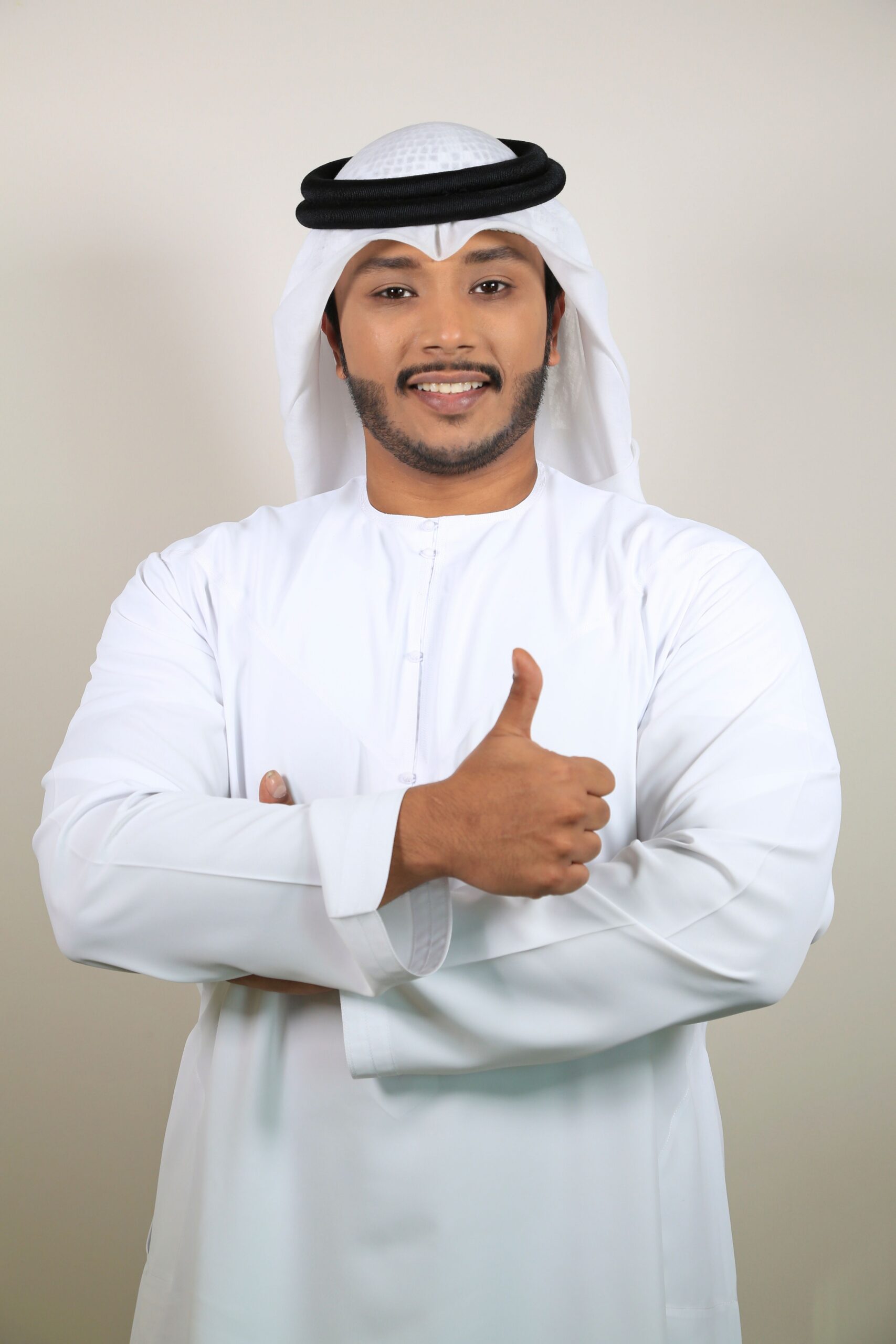 Portrait of a smiling man in traditional Emirati clothing giving a thumbs up against a white background.